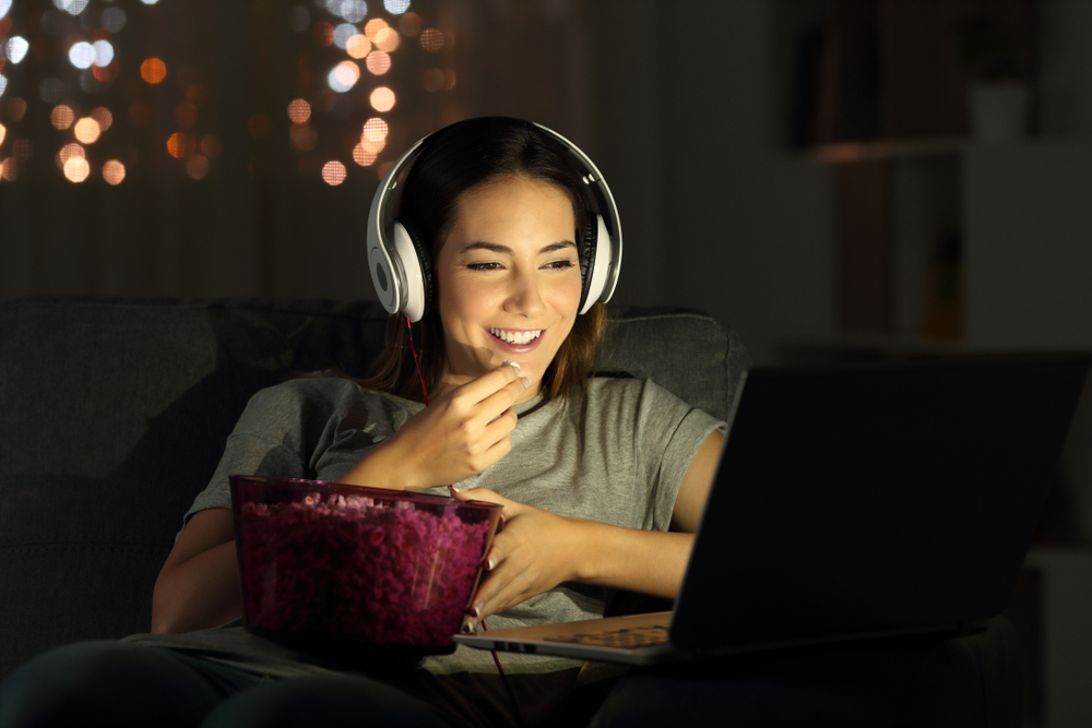 kvinna streamar film och äter popcorn framför dator med hörlurar på